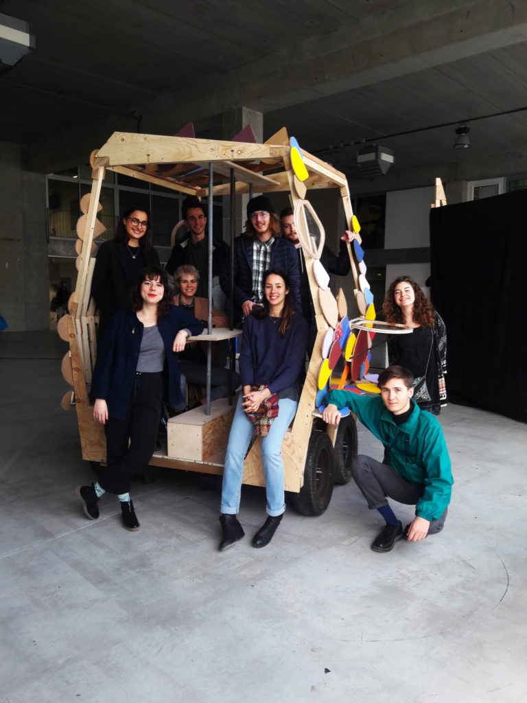 Les étudiant·es architectes posent dans le premier prototype à l'échelle d'un maillon du serpent, petite voiture en bois avec des décorations colorées faites de différents matériaux.